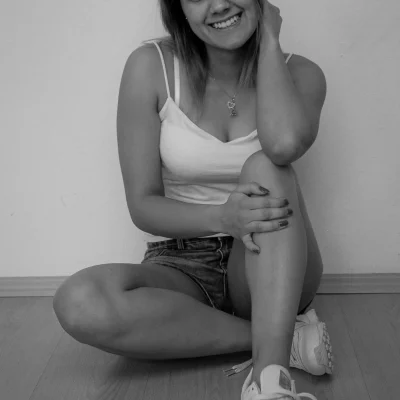 Frau mit langen Haaren, die in sommerlicher Kleidung lächelnd am Boden sitzt und ein Bein anzieht, Schnürsenkel am Sneaker offen