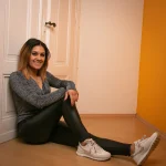 Portraitfotografie einer Frau mit langen Haaren, die lächelnd in sportlicher Kleidung auf dem Boden vor einer weißen Tür sitzt.