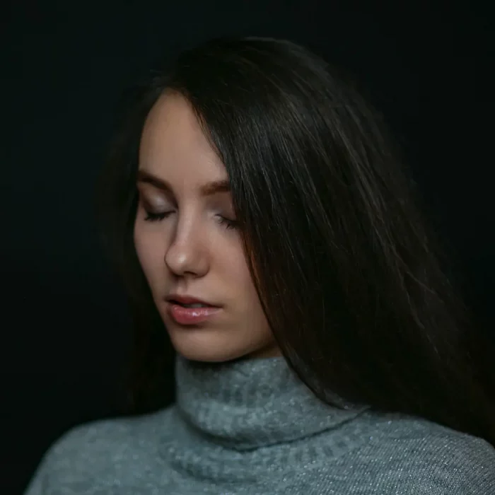 Portraitfotografie eines weiblichen Models mit geschlossenem Blick und langem Haar, frontal vor dunklem Hintergrund, grauer Rollkragenpullover