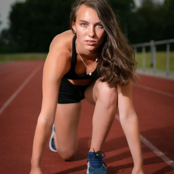 Sportplatz Fotoshooting: Model Jennifer mit langem Haar im Sportoutfit in Startposition auf roter Laufbahn, direkter Blick in die Kamera