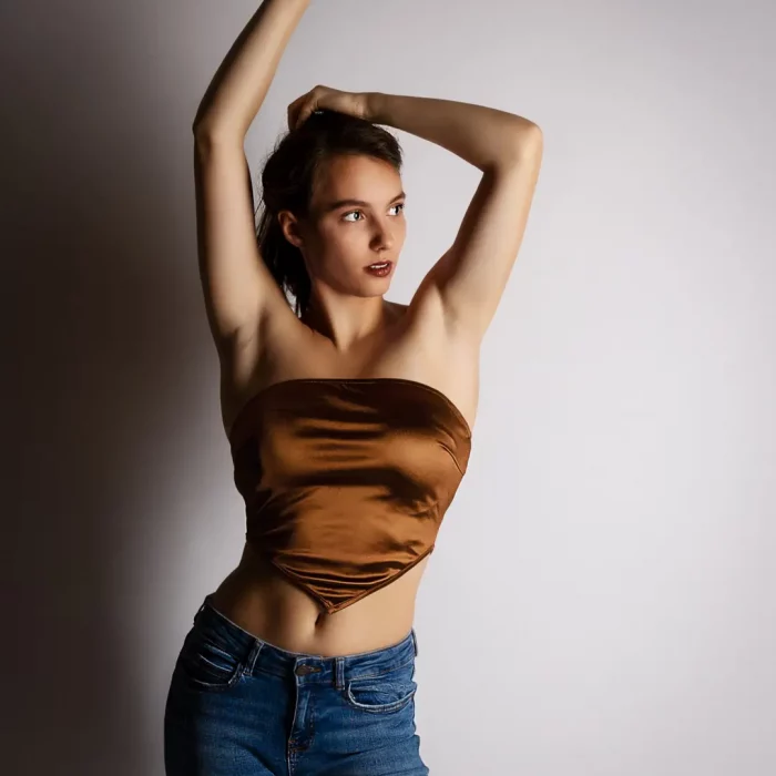 Weibliches Model mit goldenem Top und blauen Jeans posiert vor hellem Hintergrund mit erhobenen Armen.