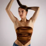 Junge Frau mit goldenem schulterfreiem Crop-Top und blauer Jeans hält ihre Haare hoch, Blick zur Seite, neutraler Hintergrund