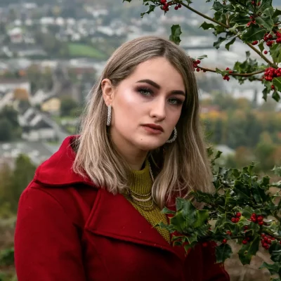 Junge Frau mit langen, blonden Haaren und rotem Mantel neben einem Beerenstrauch mit roten Beeren, unscharfe Stadt im Hintergrund