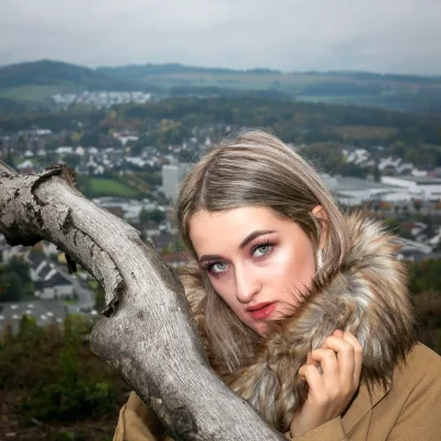 Junge Frau mit langen, hellblonden Haaren und Kunstpelzkragen, die vor einer bewaldeten Landschaft mit Stadt im Hintergrund posiert