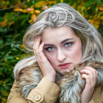 Porträtfotografie einer jungen blonden Frau mit Fellkragen vor herbstlichem Waldhintergrund, direkte Blickrichtung in die Kamera