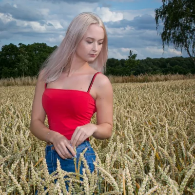 Junge Frau mit langen, hellblonden Haaren in rotem Top und Jeans steht in sommerlichem Kornfeld, Blick zur Seite