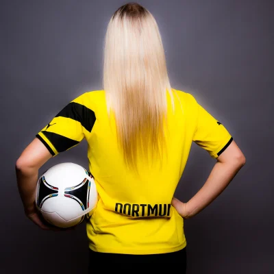 Frau mit langen blonden Haaren steht mit dem Rücken zur Kamera, trägt ein gelbes Trikot mit schwarzem Schriftzug "DORTMUND" und hält einen Fußball unter dem linken Arm
