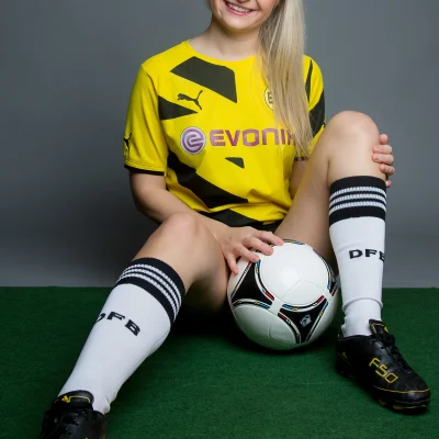 unge Frau sitzt auf grünem Teppich mit Fußball zwischen den Beinen und trägt gelbes Fußballtrikot, weiße Stutzen mit schwarzem Schriftzug und schwarze Fußballschuhe