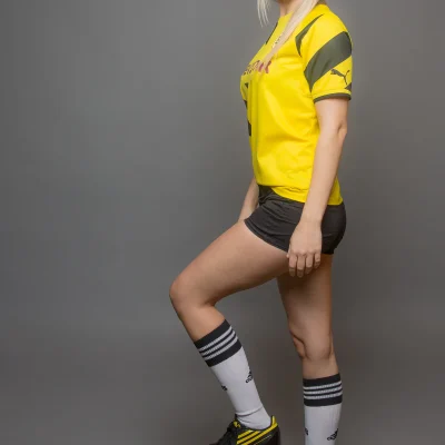 Jessica in gelbem Sporttrikot, schwarzen Shorts, weißen Kniestrümpfen und schwarzen Sportschuhen steht mit einem Fuß auf einem Fußball vor grauem Hintergrund