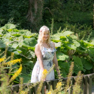Frau mit langem blondem Haar und weißem Kleid steht lächelnd auf einer Brücke vor großblättrigen Pflanzen