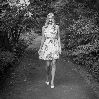 Model Jessica in floralem Kleid geht auf einem von Büschen gesäumten Weg und lächelt