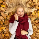 Herbst Fotoshooting in Soest: Blondes weibliches Model mit rotem Schal, das lächelnd auf gelb-braunen Blättern liegt