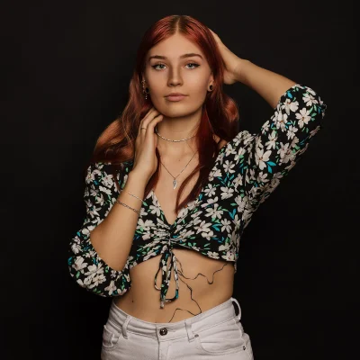 Portraitfotografie von Johanna Hellmann mit rötlich-braunen Haaren, geblümter Bluse und weißer Hose vor schwarzem Hintergrund, eine Hand im Haar