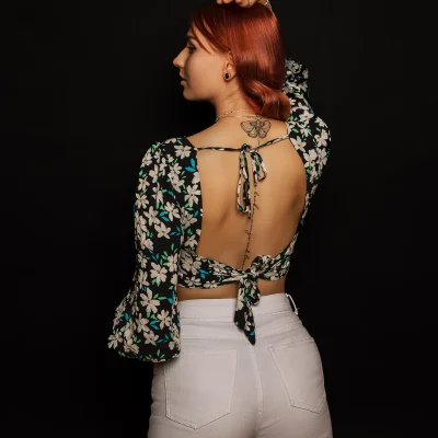 Johanna mit rötlichen Haaren, floraler Bluse mit offenem Rücken und sichtbarem Schmetterlingstattoo, vor schwarzem Hintergrund