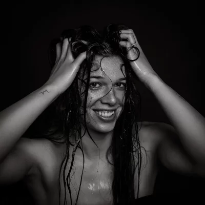 Schwarz-weiß-Portrait einer Frau mit nassem Haar und nassem Gesicht, die die Hände in den Haaren hält, mit einem kleinen Tattoo am linken Unterarm und einem Ring am rechten Ringfinge
