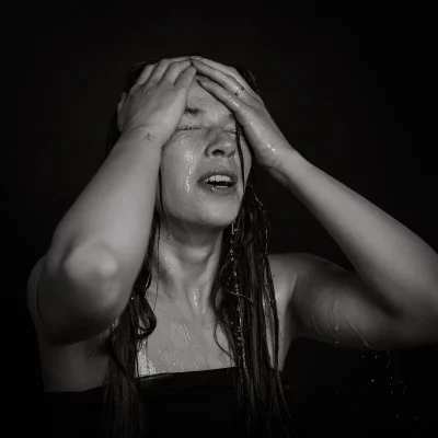 Frau mit nassen Haaren hält sich mit beiden Händen den Kopf vor schwarzem Hintergrund, Wasser tropft von Haut und Haaren - TFP-Fotoshooting
