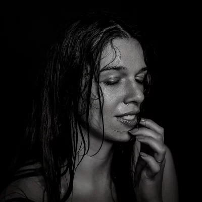 Model Julia mit nassen Haaren vor schwarzem Hintergrund, die sich mit der rechten Hand an das Gesicht fasst. Im Gesicht sind Wassertropfen.