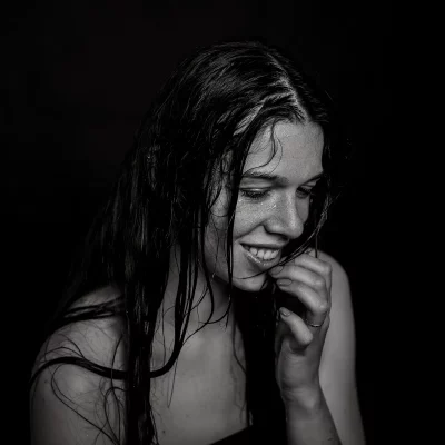 Frau mit nassen, langen Haaren, die sich mit der Hand an das Kinn fasst vor schwarzem Hintergrund. Wasser ist im Gesicht.