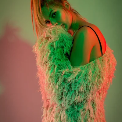 Frau in einer fransigen Jacke sitzt seitlich auf einer Stuhllehne, grüne und rötliche Farbfilter betonen Gesicht und Kleidung.