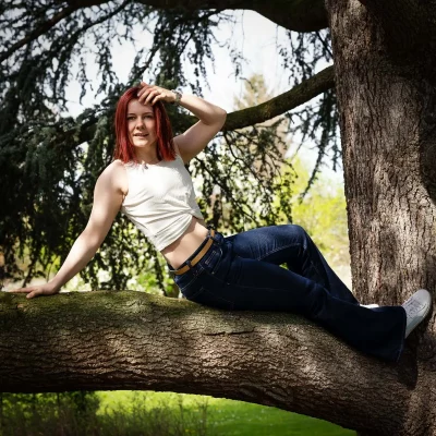 Frau mit rotem Haar in weißem ärmellosem Oberteil und blauer Jeans liegt auf einem dicken Baumast in Soest