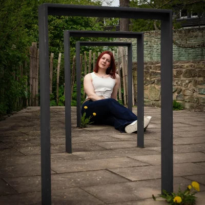 Frau sitzt auf dem Boden hinter mehreren rechteckigen Metallrahmen in einem Gartenweg mit Steinmauer im Hintergrund