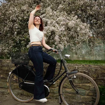 Model Katarina in weißem ärmellosem Oberteil und dunkler Hose sitzt auf einem schwarzen Fahrrad vor blühendem Baum und hebt eine Hand