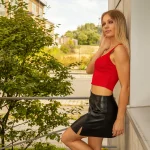 Kathleen Unger mit rotem Top und schwarzem Lederrock, an eine Wand gelehnt, Hintergrund Natur und moderne Architektur