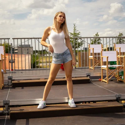 Junge Frau in lässiger Sommermode, weißes Top und Jeansshorts, steht selbstbewusst auf Tribüne vor Sitzreihen im Freien.