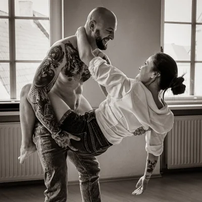 Portraitfotografie eines tätowierten, oberkörperfreien Mannes, der eine lachende Frau im Arm hält, im Innenraum vor großen Fenstern