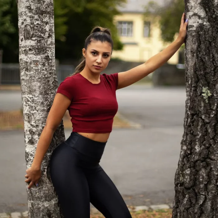 Fotoshooting mit Kim Soest: Junge Frau mit dunklen Haaren in rotem Oberteil und schwarzen Leggings steht zwischen zwei Baumstämmen im Freien