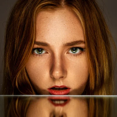 Portraitfotografie einer jungen Frau mit roten Haaren, die ihr Gesicht nah über einen Spiegel hält und deren Spiegelbild sichtbar ist