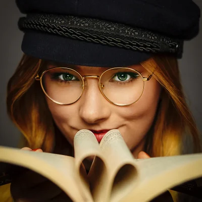 Junge Frau mit goldener Brille und schwarzer Mütze, die ein aufgeschlagenes Buch in den Händen hält.