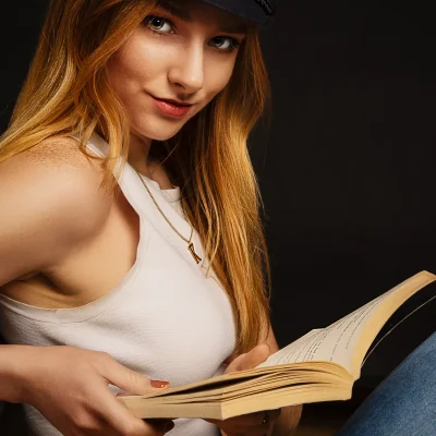 Frau mit langen Haaren, weißem Oberteil und Mütze, die ein aufgeschlagenes Buch hält und direkt in die Kamera lächelt.