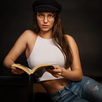 Eine Frau mit Brille und dunkler Mütze, die in weißem Top und Jeans ein aufgeschlagenes Buch hält und in die Kamera blickt.