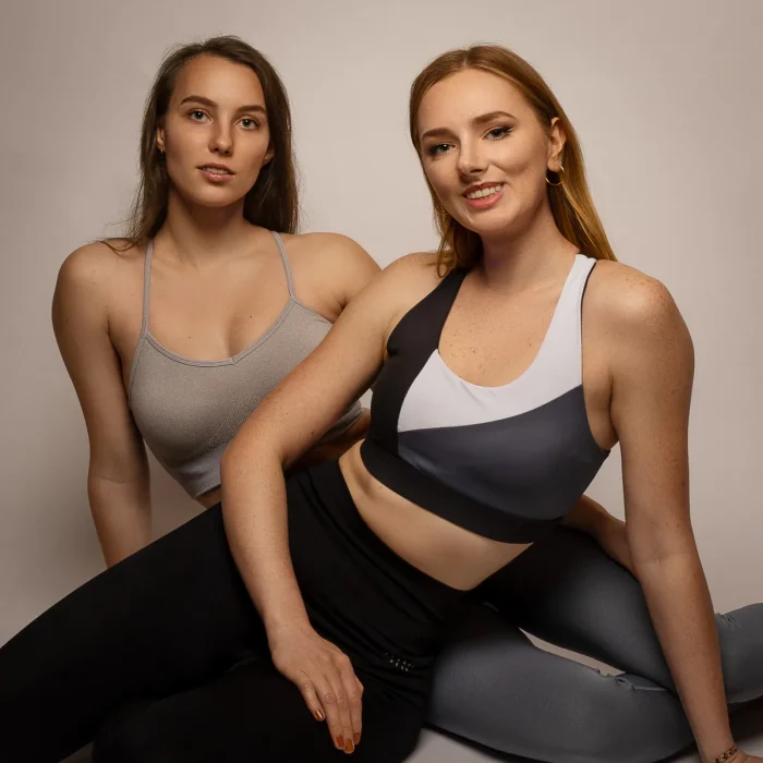 Freundinnen Fotoshooting: Zwei Frauen in Sportkleidung sitzen nebeneinander vor einem hellen Hintergrund