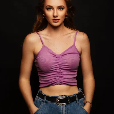 Klaudia mit hochgestecktem Haar, lila ärmellosem Top mit Raffung vorne und blauer Jeans vor schwarzem Hintergrund