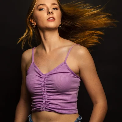 Frau mit langen, rotblonden, seitlich wehenden Haaren, in lila Top und Jeans, vor dunklem Hintergrund
