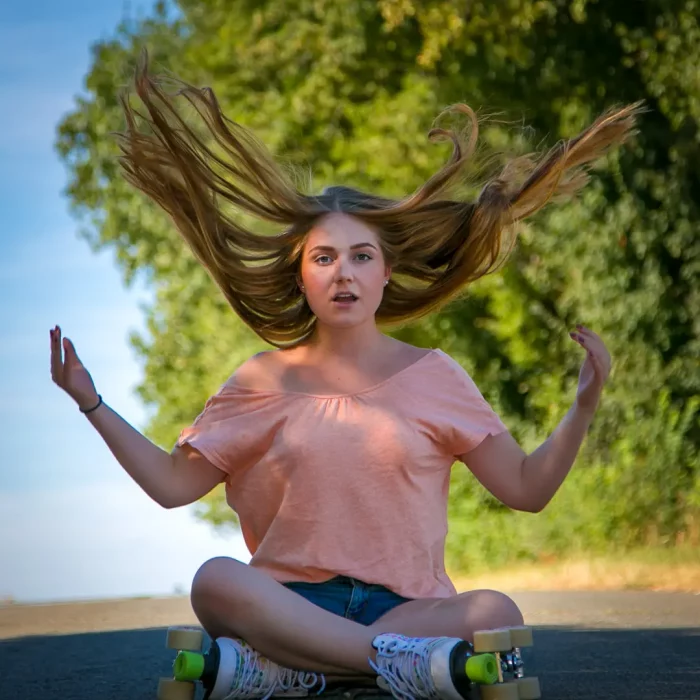 Frau mit langen, hochgeschleuderten Haaren sitzt im Schneidersitz auf Rollerskates auf einer Straße.