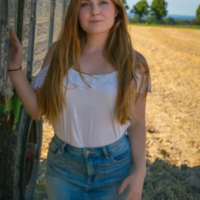 Frau steht im Sonnenlicht neben einem hölzernen Heuwagen auf einem abgeernteten Feld, trägt weißes Top und Jeansrock.