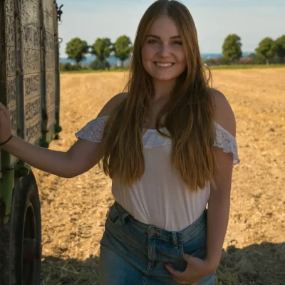 Frau mit langen, offenen Haaren steht in sommerlicher Kleidung neben einem Heuwagen auf einem Feld.