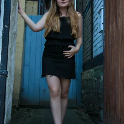 Frau mit langen Haaren in einem schwarzen Kleid, barfuß stehend in einer engen Gasse vor einer blauen Holztür