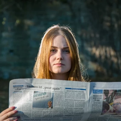 Junge Frau mit langen, blonden Haaren, die bei Tageslicht eine aufgeschlagene Zeitung hält und direkt in die Kamera blickt