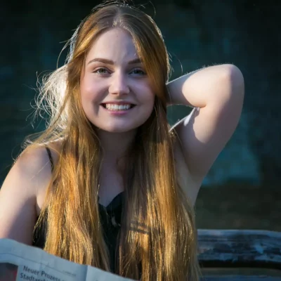 Porträtfotografie einer jungen Frau mit langen, blonden Haaren, die auf einer Bank sitzt und eine Zeitung hält.