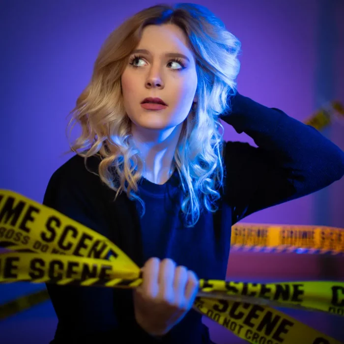 Blonde Frau mit nachdenklichem Blick hält gelbes Crime-Scene-Absperrband vor lilafarbenem Studiohintergrund.