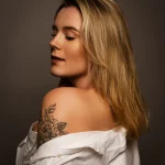 Frau mit aufgeknöpfter weißer Bluse und floralem Schulter-Tattoo, seitliches Profil vor neutralem Hintergrund