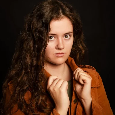 Lena in cognacfarbener Wildlederjacke zieht den Kragen hoch, ernste Mimik, direkter Blick – Schauspielportrait vor schwarzem Hintergrund