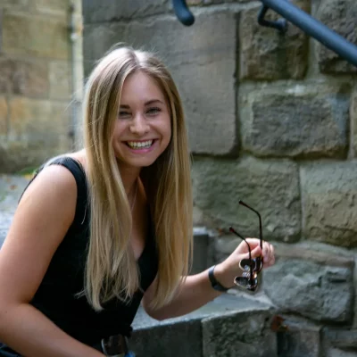 Junge Frau mit langen, blonden Haaren, die im Sitzen auf einer Steintreppe lacht und eine Sonnenbrille in der Hand hält