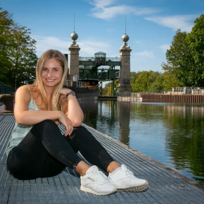 Lisa-Carolina, sitzend auf einem Steg vor dem historischen Schiffshebewerk, inmitten grüner Bäume und blauem Wasser