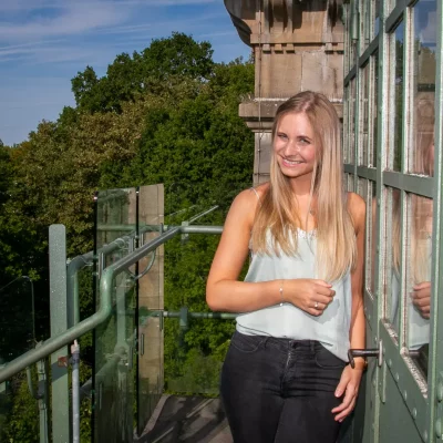 Porträtfotografie einer jungen Frau mit langen blonden Haaren, die lächelnd auf einer Brücke mit grüner Verglasung steht.