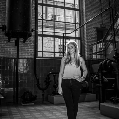 Frau posiert im schwarz-weißen Porträt vor gemauerten Wänden und großen Industriefenstern in einer alten Fabrikhalle.